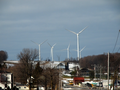 [Houses with trees around them in the foreground with five windmills on the hill above them. One is much closer and thus larger than the other four. The blades are turning and thus each in different positions.]